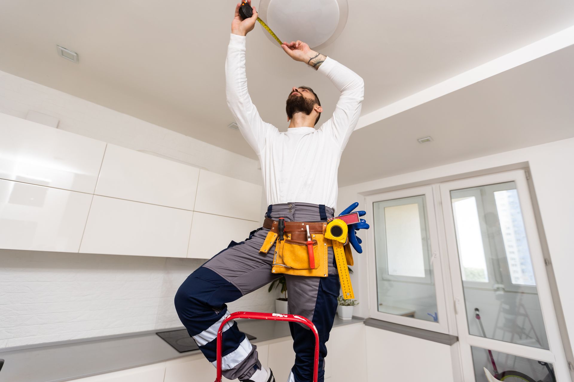 An electrician on a ladder installing a ceiling light in a modern kitchen.