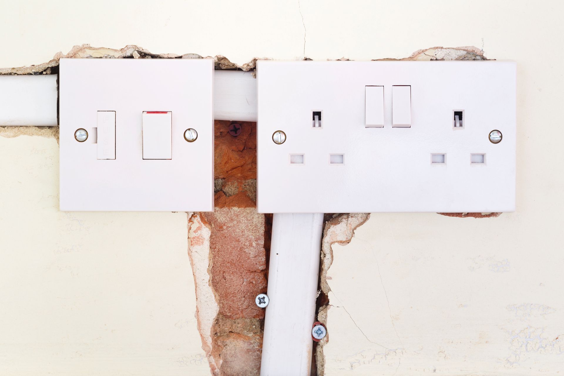 White light switches and electrical outlets on a damaged wall with exposed brick and wiring.