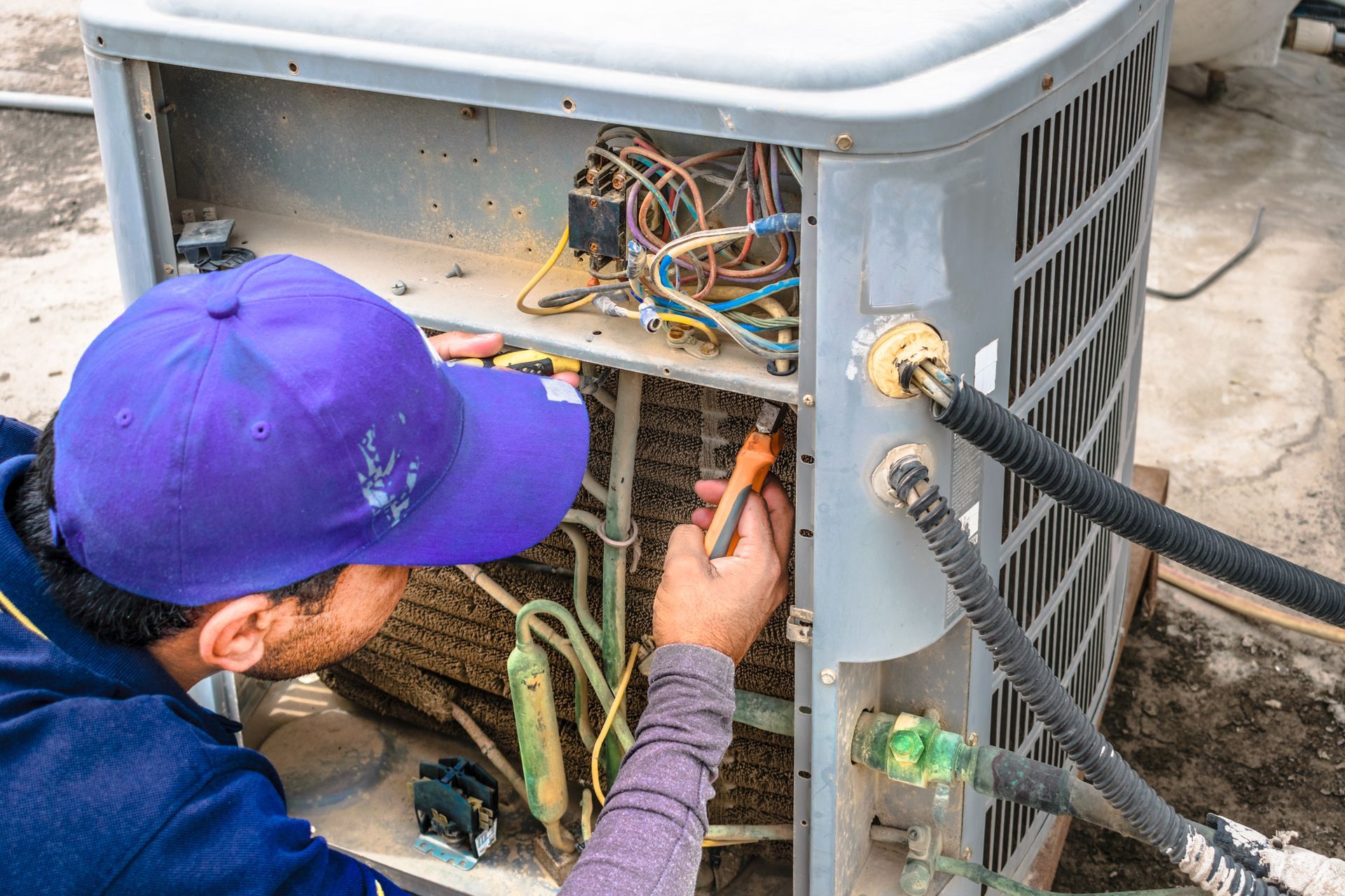 HVAC technician in blue cap repairs an air conditioning unit, using tools.