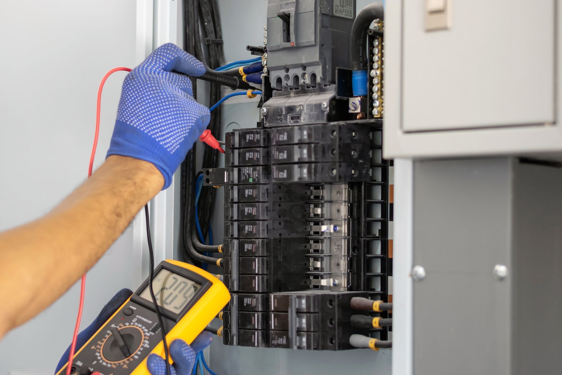 An electrician in blue gloves is using a multimeter to test a circuit breaker panel.