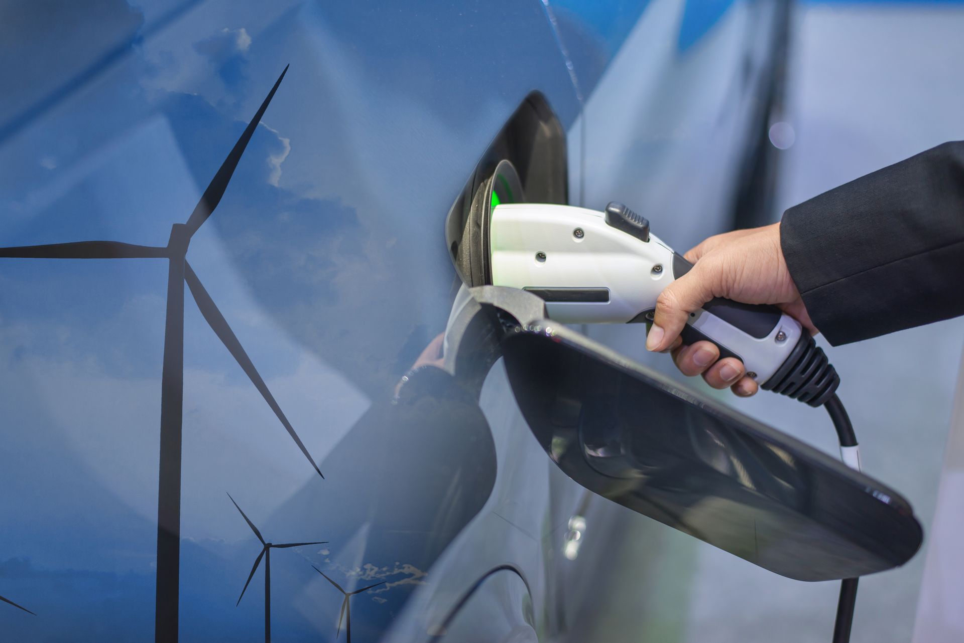 Person charging an electric car, using a charging cable; background shows wind turbines.