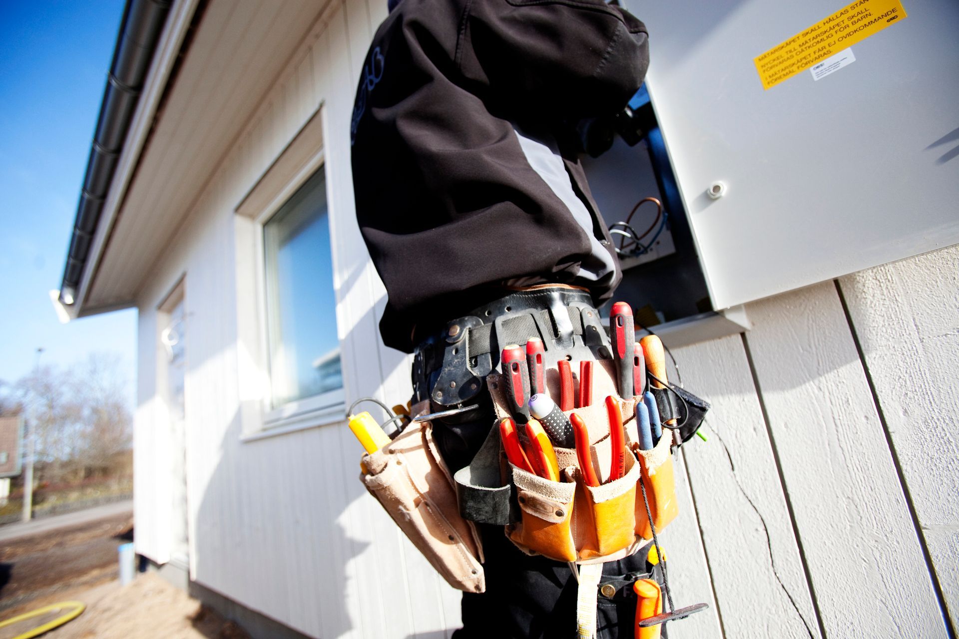 Electrician working on an electrical box, wearing a tool belt, next to a white house.