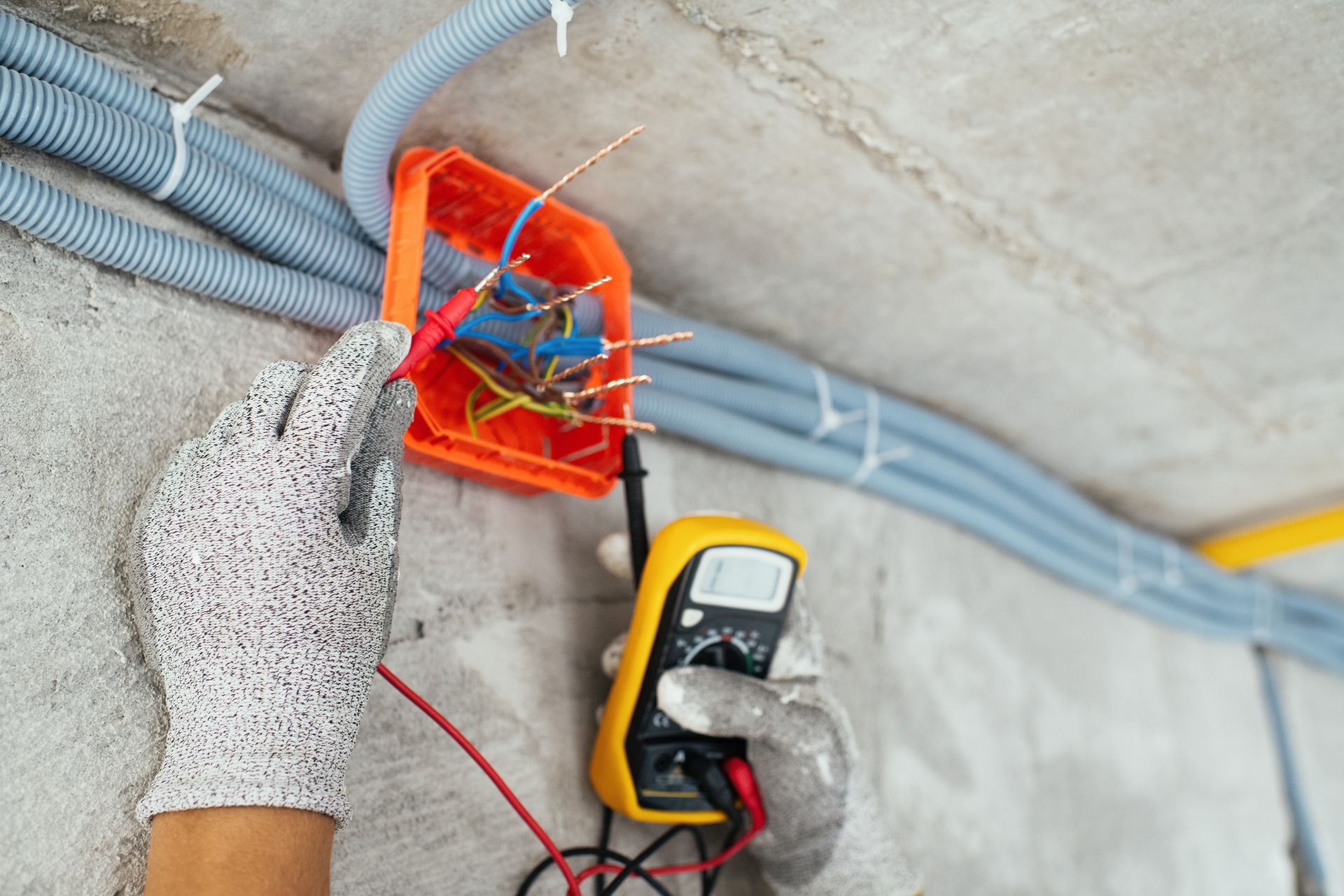 Electrician testing wiring with a multimeter in an orange junction box, wearing gloves, on a concrete wall.