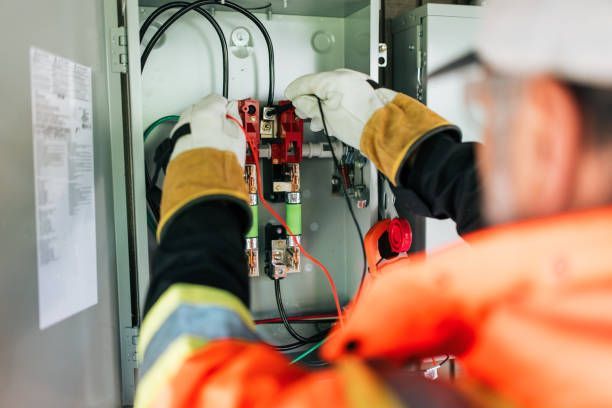 An electrician in an orange vest and gloves is working inside an electrical panel.