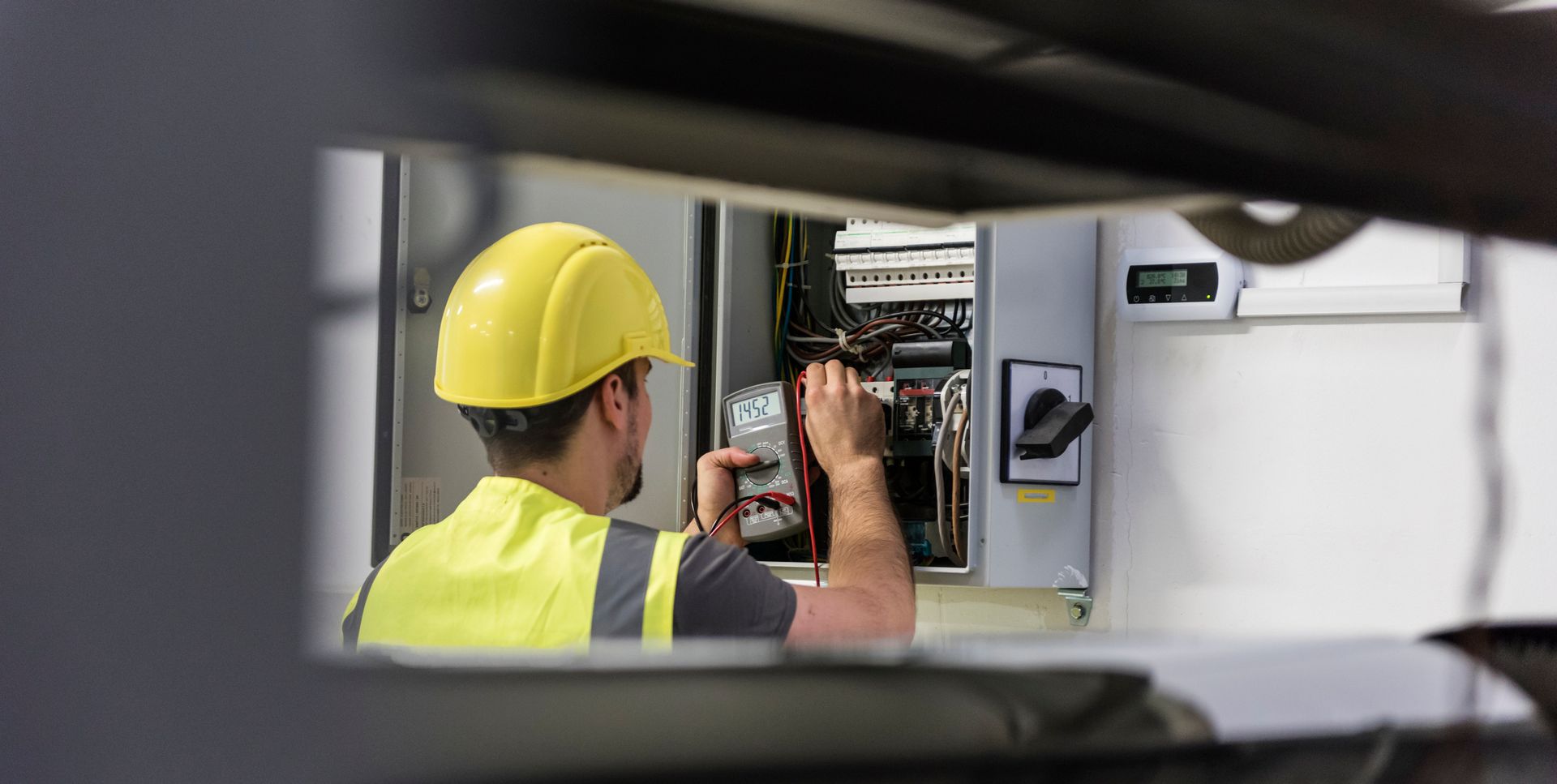 An electrician wearing a yellow hard hat and safety vest works on an electrical panel.