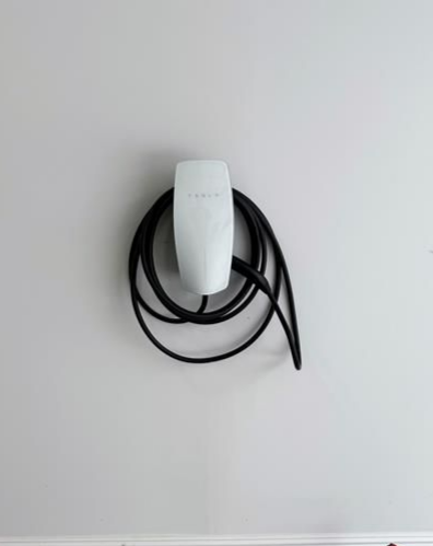 White Tesla EV charger mounted on a light gray wall with black charging cable coiled around it.