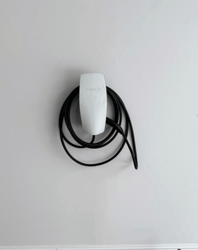 White Tesla EV charger mounted on a light gray wall with black charging cable coiled around it.