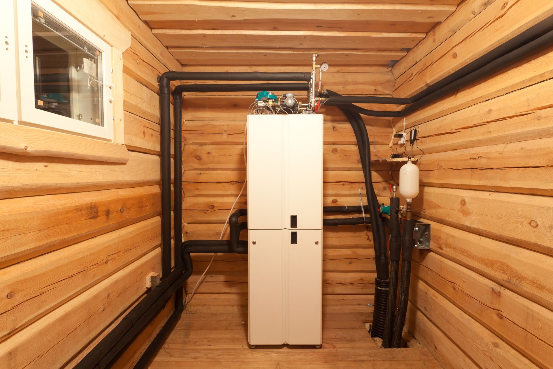 White utility cabinet in a wood-paneled room, surrounded by black pipes and a small window.