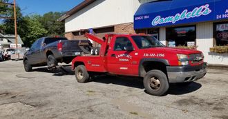 A red tow truck is towing a black truck in front of a campbell 's restaurant.