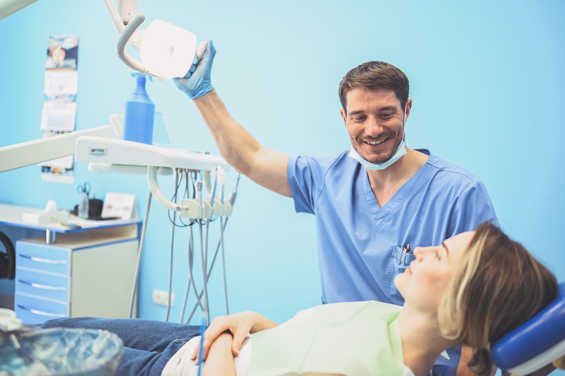 Dentist adjusting light over patient in dental chair, blue scrubs, dental office setting.