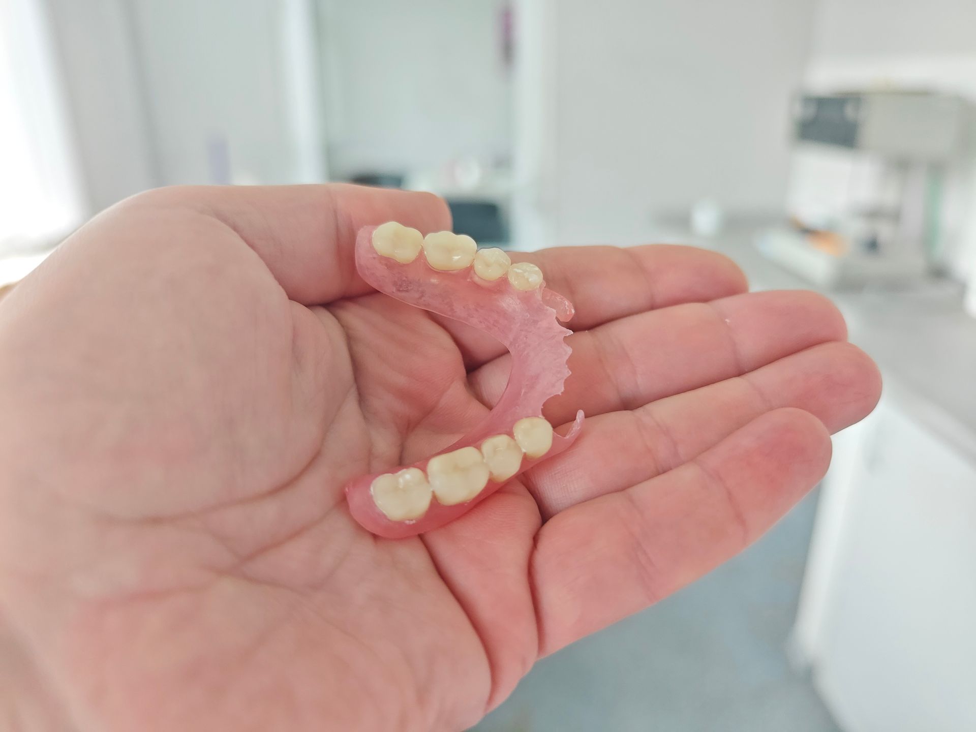 A hand holds a set of partial dentures, with a blurry office in the background.