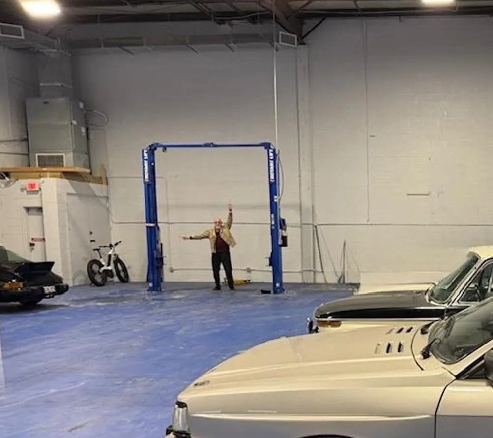 Person standing under a car lift in a garage, arms outstretched, celebrating. Classic cars are parked nearby.
