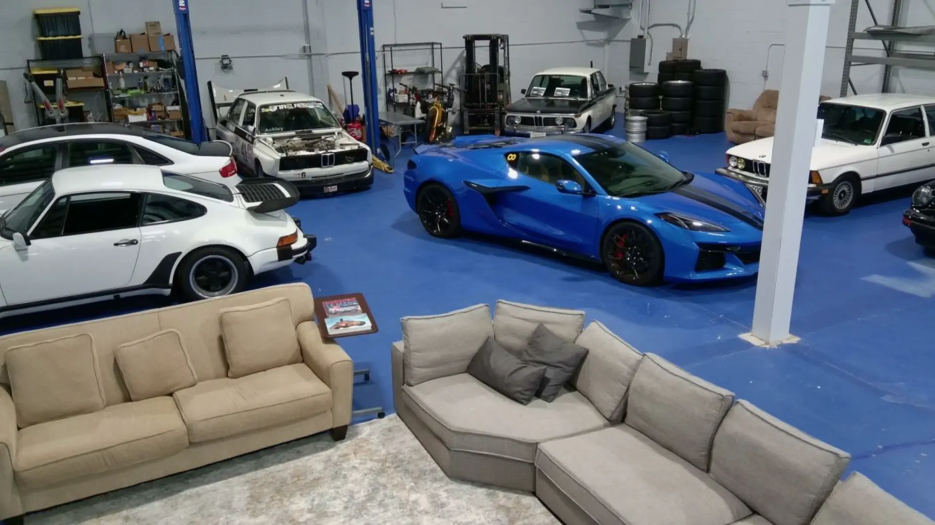 Blue Corvette and other cars in a garage setting with couches and a rug.