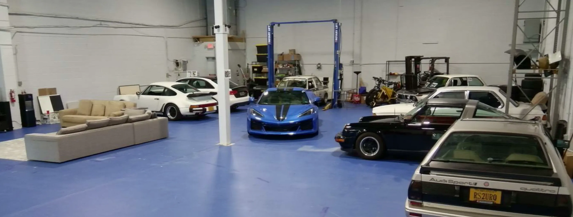 A blue sports car parked inside a garage, with other cars and furniture visible.