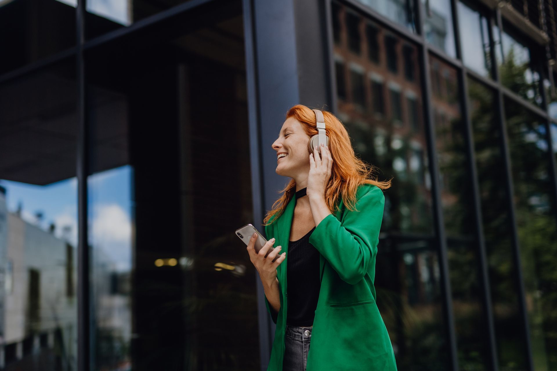 A woman in a green jacket is wearing headphones and holding a cell phone. | Brooklyn, NY | Urban Hearing