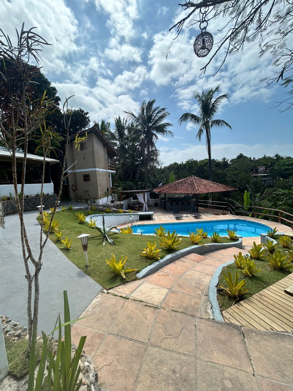 Um pátio amplo e ensolarado com piscina curva, gramado, caminho de pedra, estrutura com telhado de palha e árvores tropicais exuberantes.