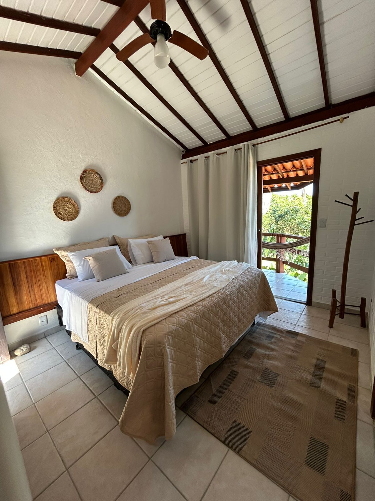 Um quarto moderno com cama queen-size, roupa de cama creme, paredes brancas, vigas de madeira expostas no teto e vista para uma varanda externa.