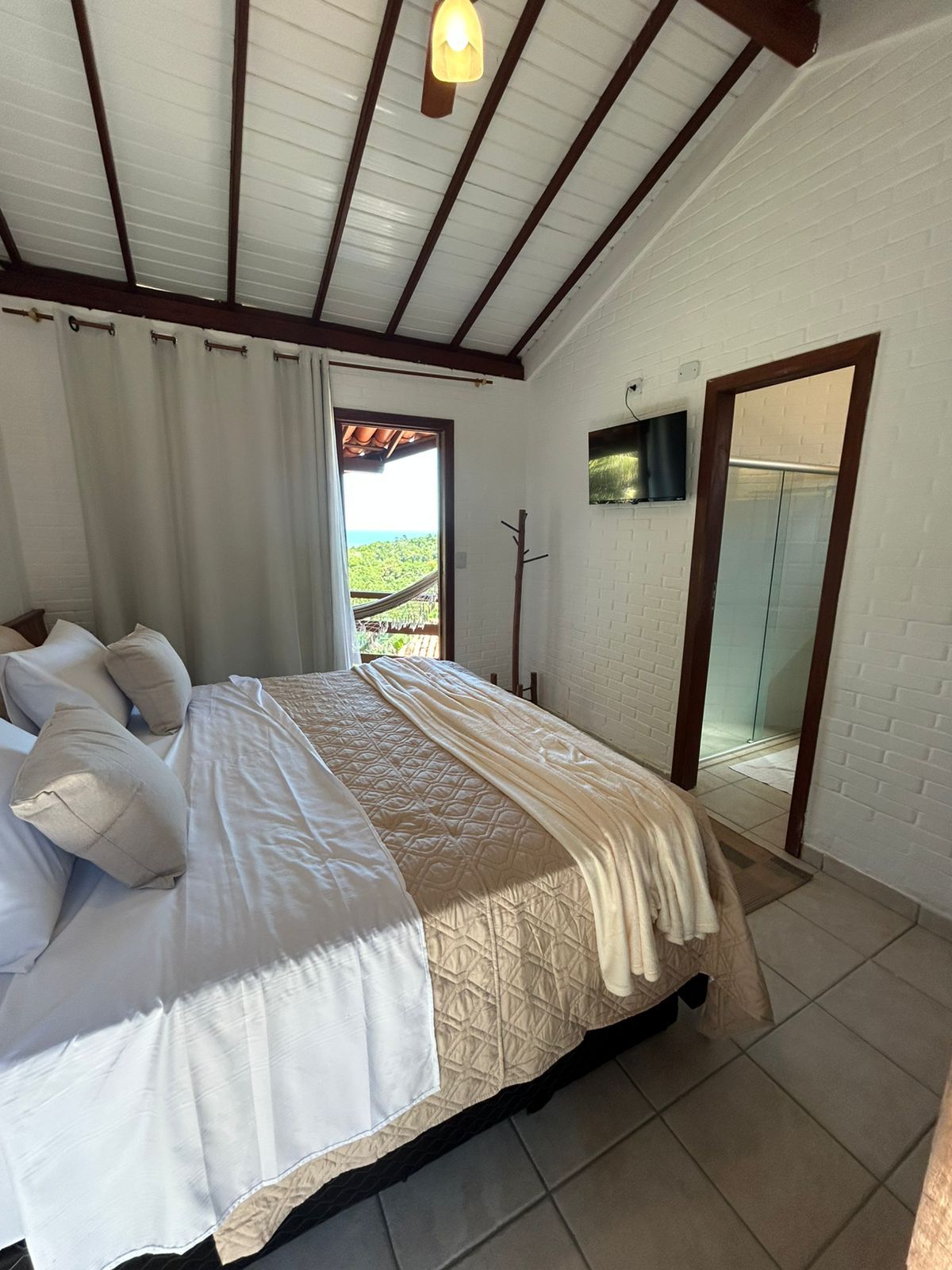 Um quarto de hotel com cama arrumada, teto com vigas de madeira, vista para uma varanda e uma porta aberta que dá acesso a um banheiro.