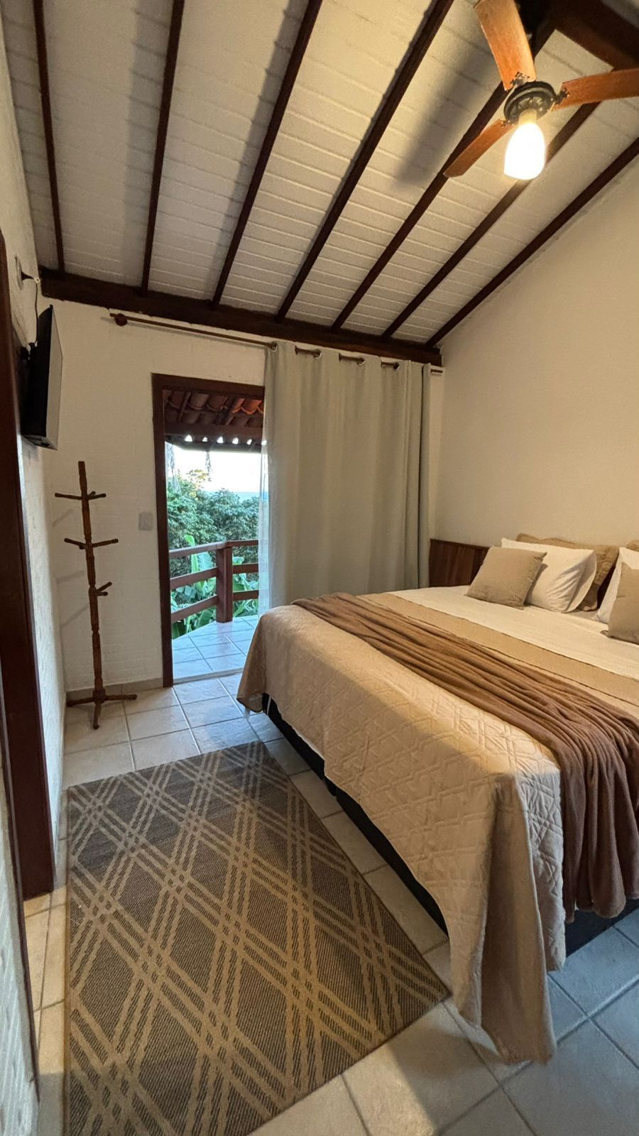 Um quarto com cama, tapete com estampa de losangos, cabideiro de madeira, ventilador de teto e porta que dá para uma varanda.