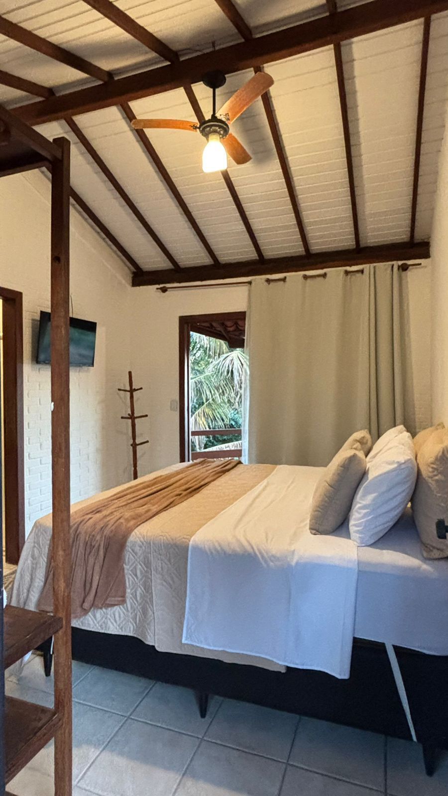 Um quarto arrumado dispõe de uma cama de casal com lençóis e travesseiros brancos, um teto com vigas aparentes e ventilador, além de vista para a vegetação.