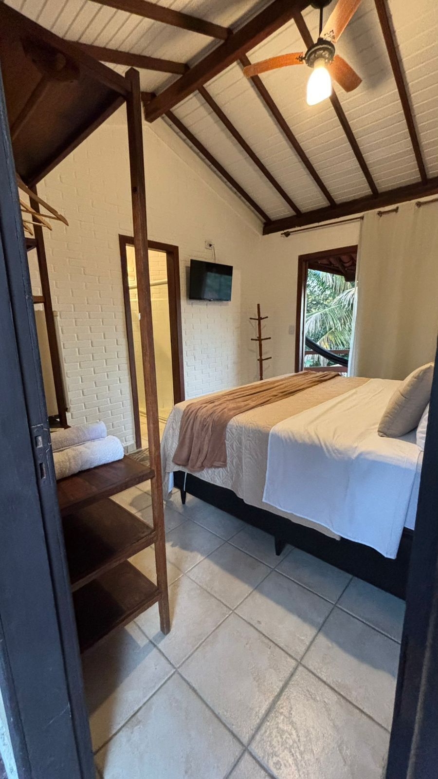 Um quarto luminoso com piso de azulejos brancos, uma cama queen-size com roupa de cama em tons neutros, vigas de madeira no teto e um banheiro aberto.