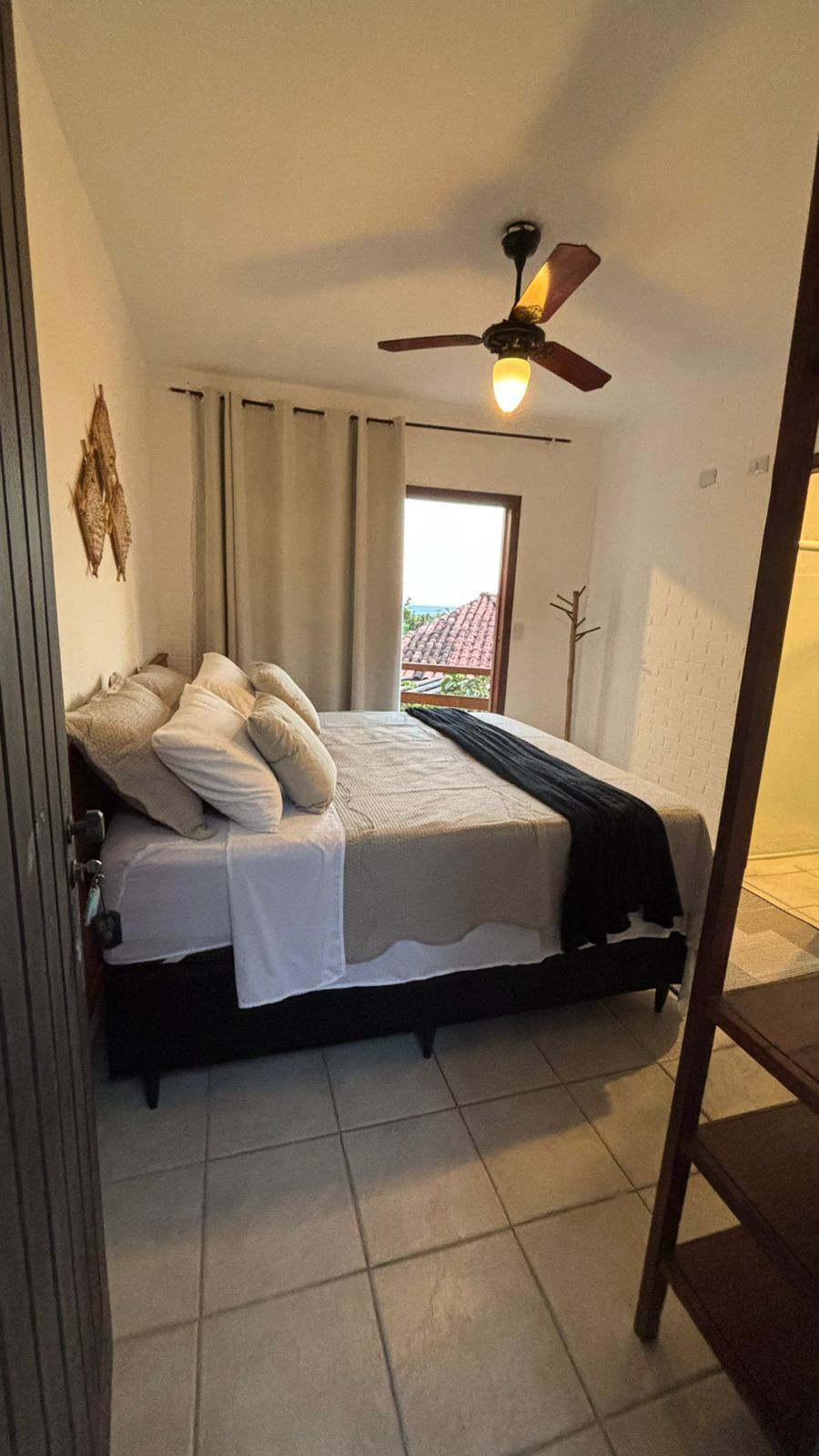 Um quarto com cama de casal com roupa de cama em tons neutros, ventilador de teto, cortinas bege e piso de azulejo claro.