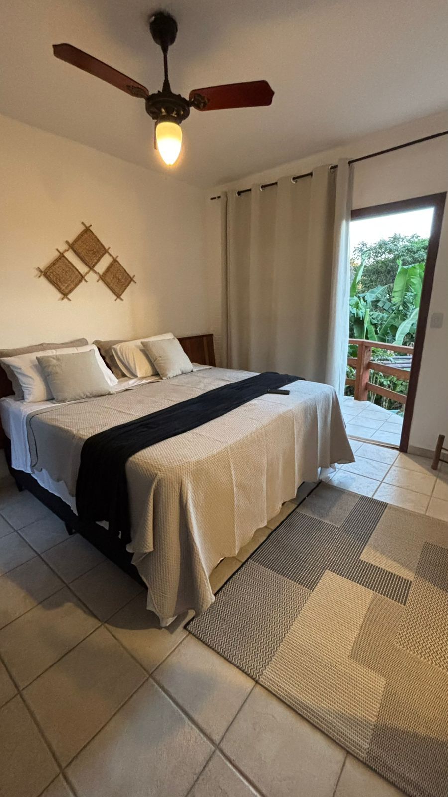 Um quarto luminoso com uma cama com um peseira preta, um tapete geométrico, um ventilador de teto e uma porta que dá para uma varanda.