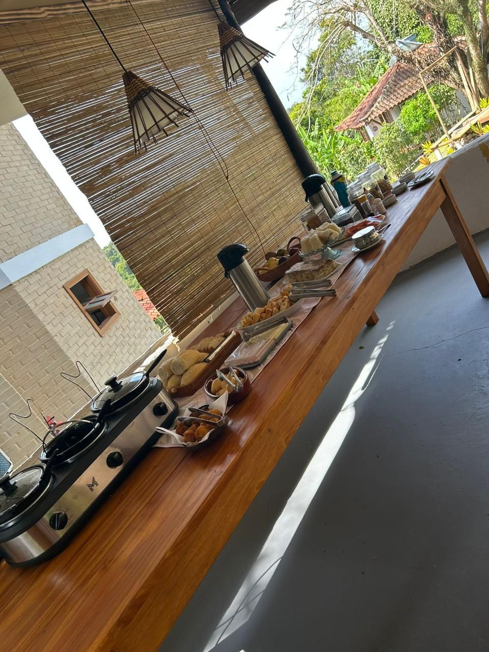 Uma longa mesa de madeira posta com comida e garrafas de café para um café da manhã em estilo bufê em uma varanda com persianas de bambu trançado.