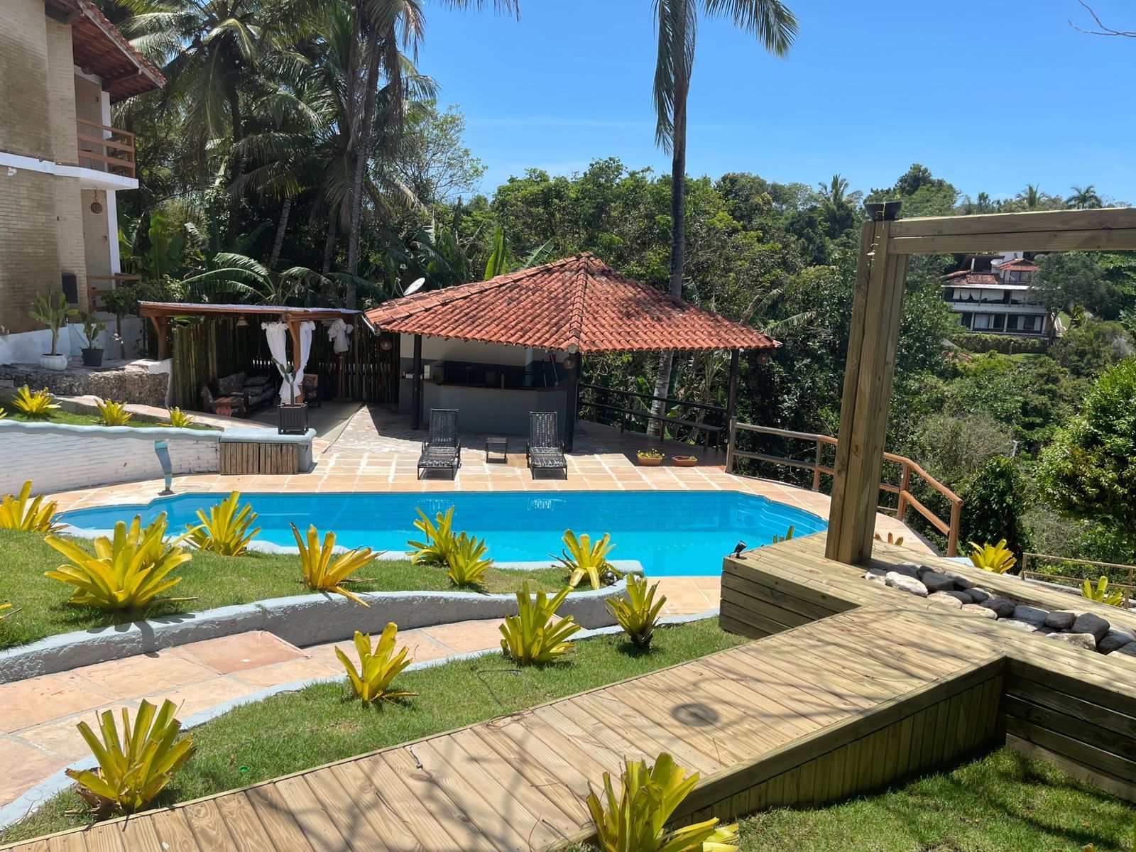 Uma área de piscina externa ensolarada com piscina azul, gazebo com teto de palha, vegetação exuberante e um deck de madeira.