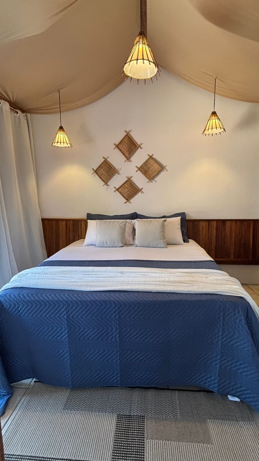 Um quarto aconchegante com uma cama de colchas azuis, travesseiros claros, painéis de madeira rústica e luminárias pendentes sob um teto de tecido.