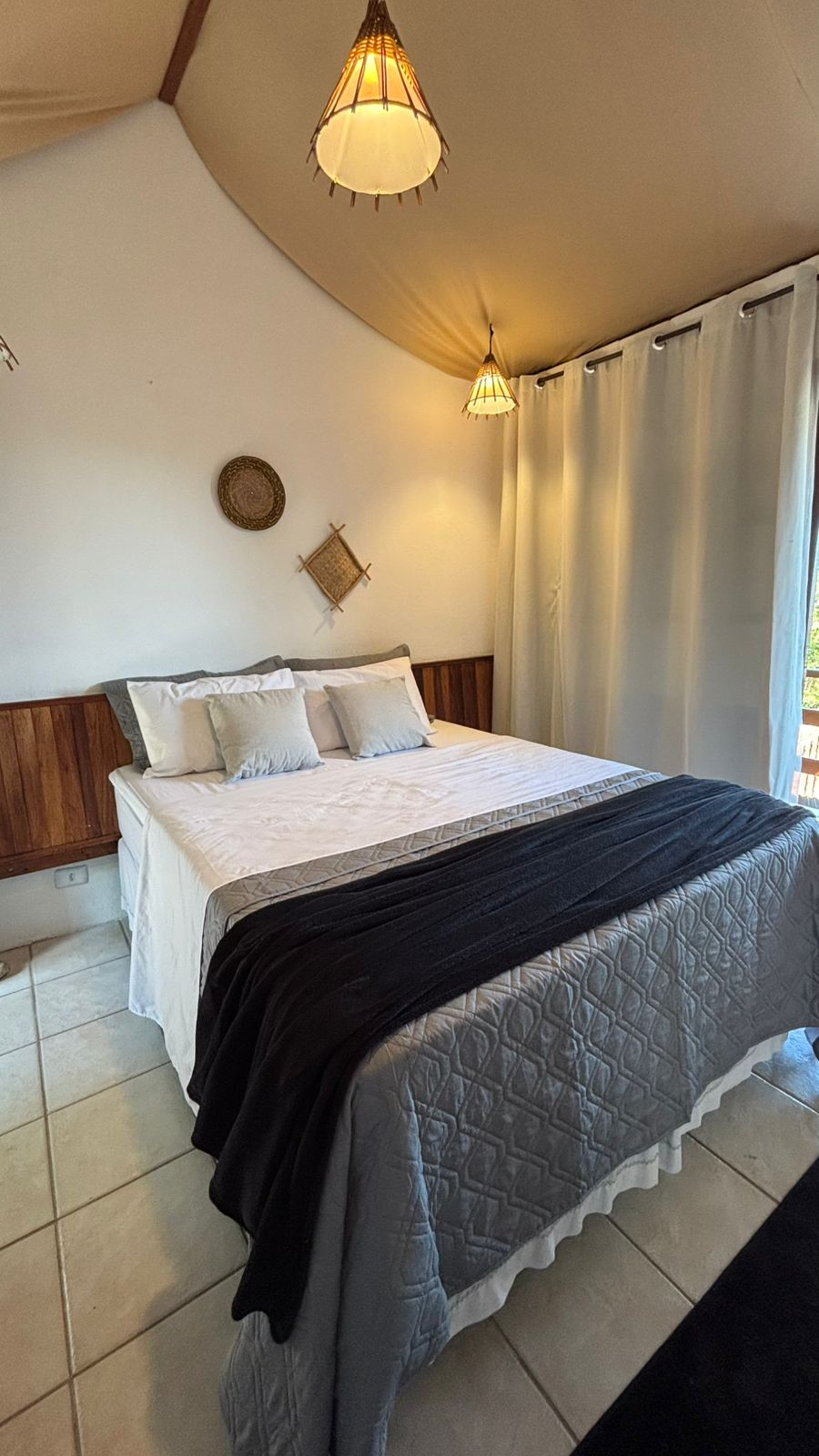 Um quarto com uma cama de casal queen-size, lençóis brancos, manta cinza, dois travesseiros e decoração de parede em tecido sob luminárias pendentes.