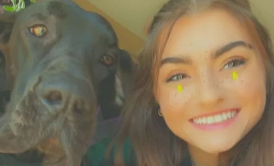 Woman with brown hair smiles, posing with a large black dog, both looking at the camera.
