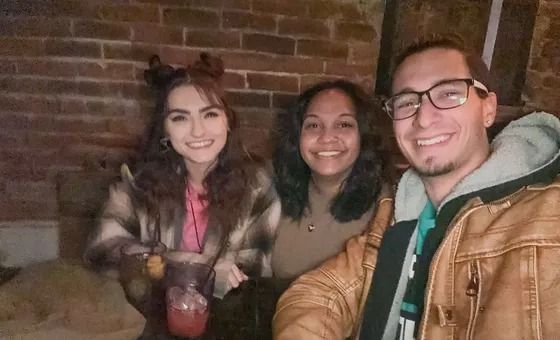 Three people smiling, seated in a dimly lit space with a brick wall. One holds a drink.