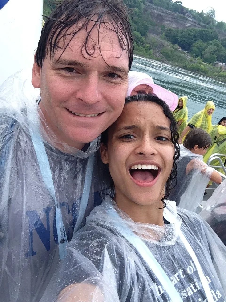 Man and young person smiling in rain ponchos, on boat tour. Water droplets, mountain background.