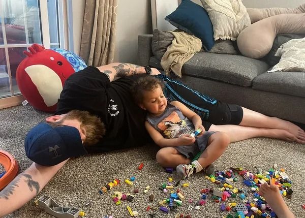 Man and child lying on carpet amidst scattered toys, near a sofa.
