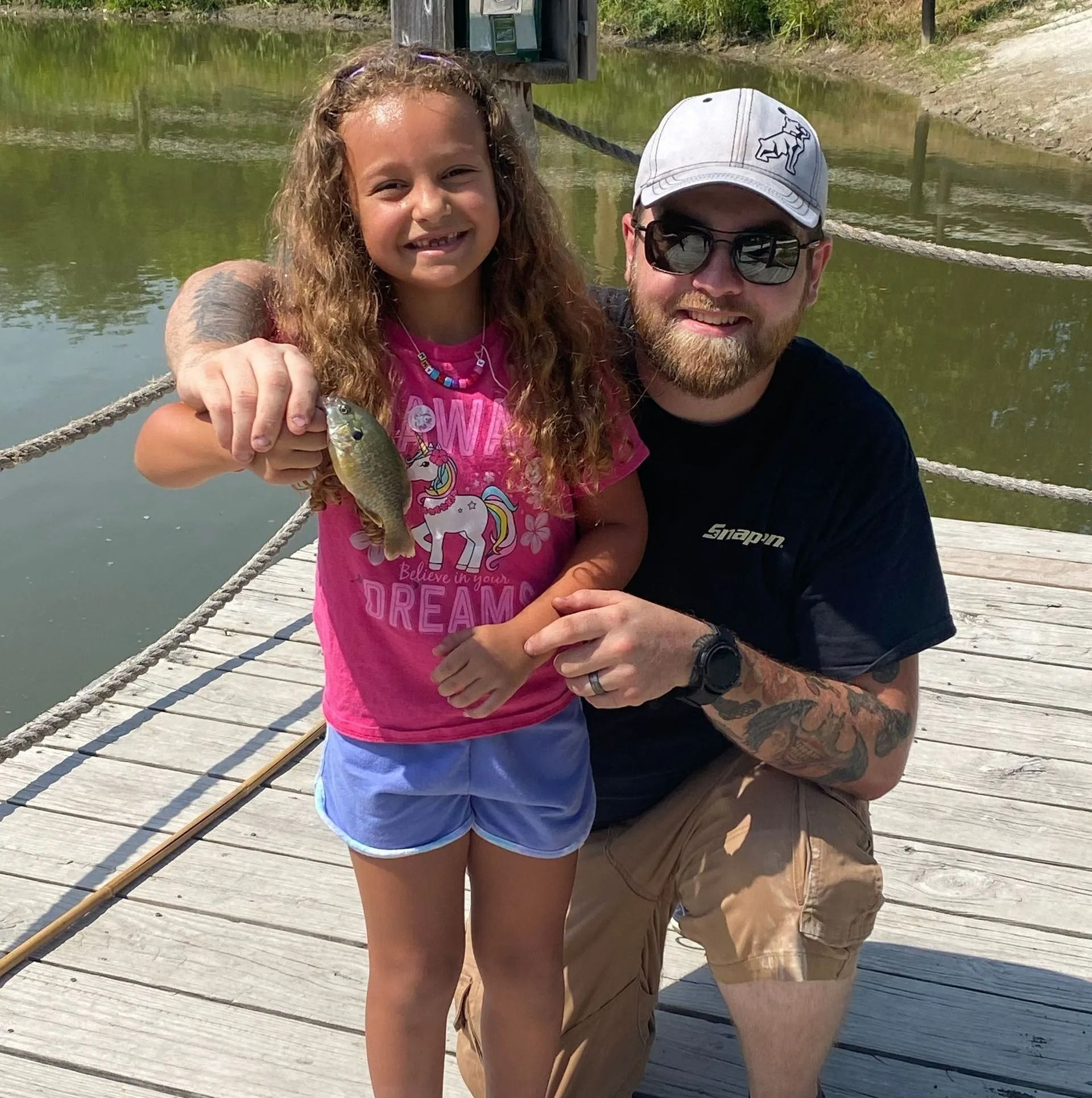 Girl and man on a dock, smiling, holding a small fish. Sunny day; girl wears pink shirt, man wears a cap.