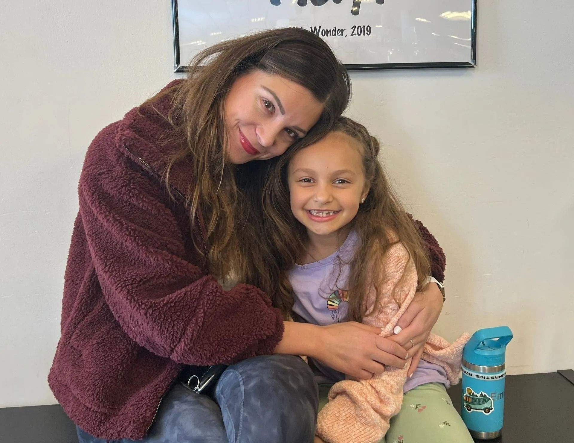 Woman and child embrace, both smiling. The woman wears a burgundy jacket, child a pink sweater.