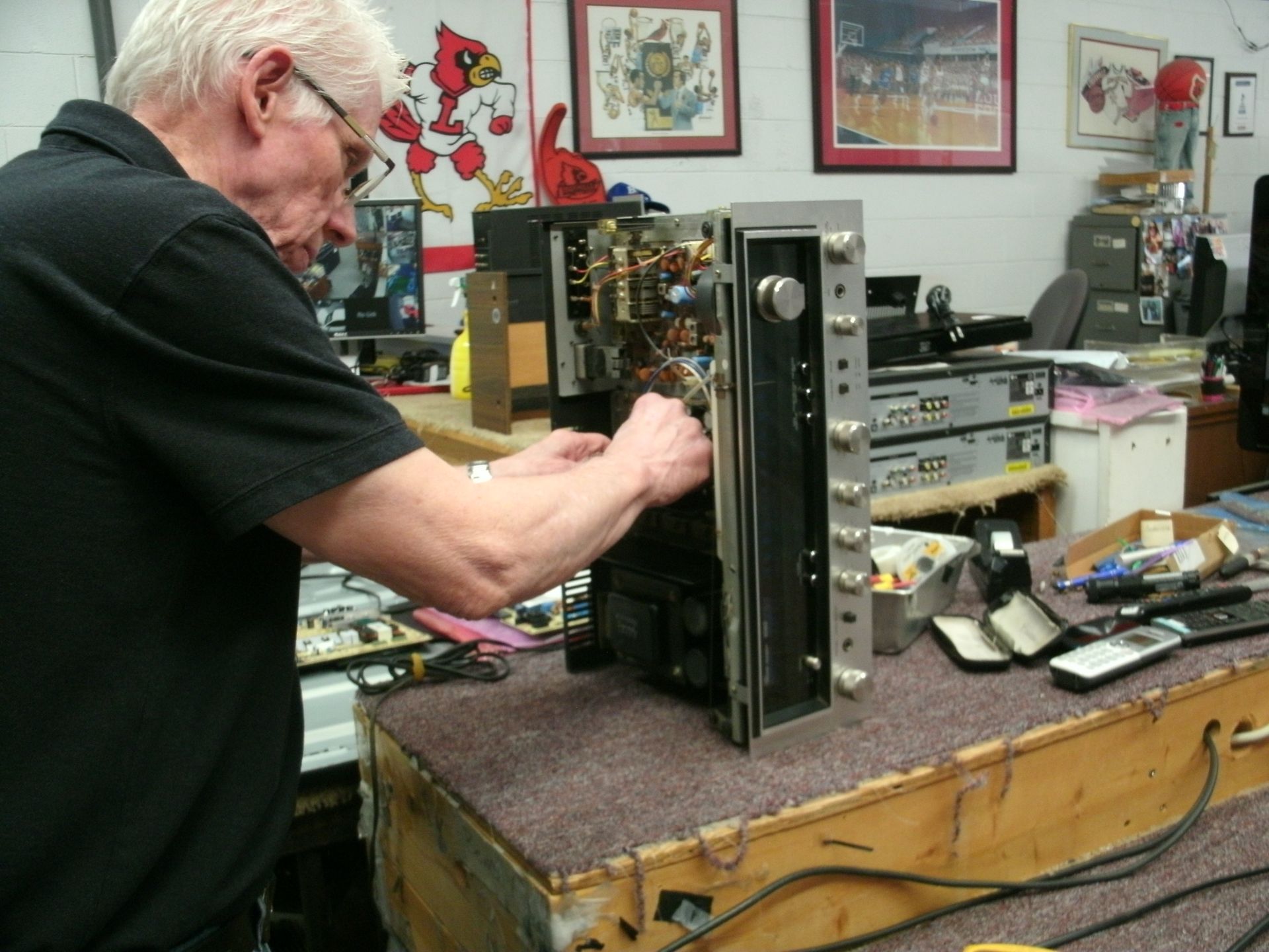 Older man repairs vintage stereo receiver on a workbench, cluttered with tools and parts.