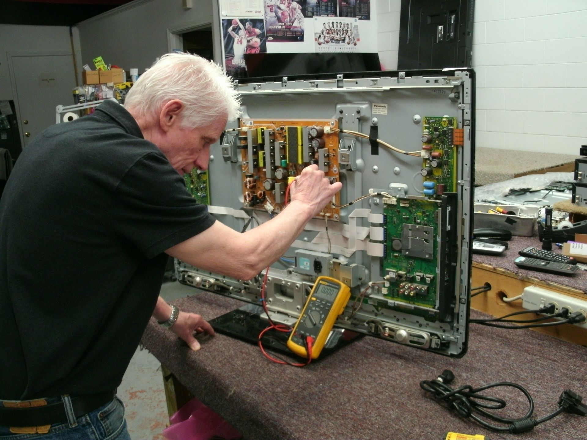 Man repairs a TV using a multimeter in a workshop. Gray hair, black shirt, electronics visible.
