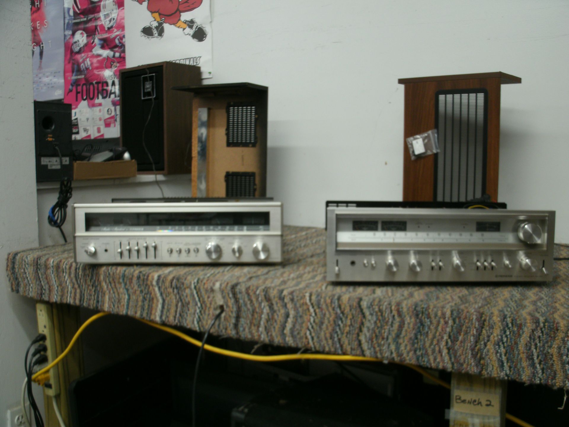 Two vintage stereo receivers sit on a patterned surface, with speakers in the background.