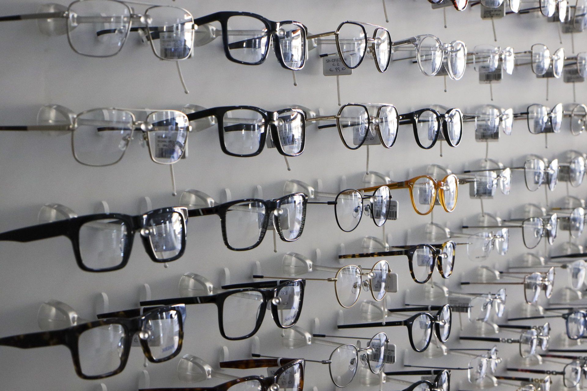 Occhiali esposti su una parete bianca in un negozio, molti con montatura nera, alcuni dorati.