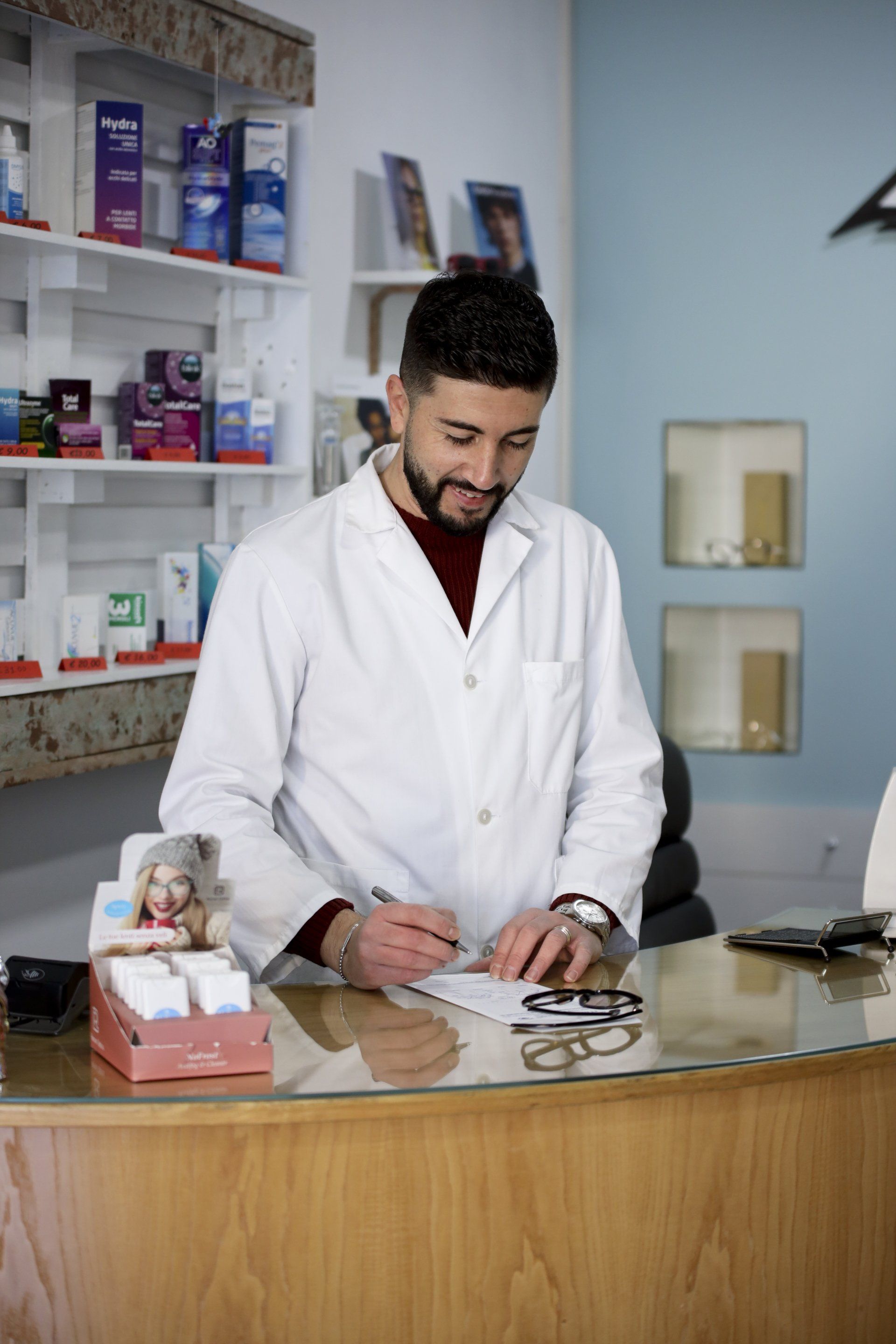 Ottico in camice bianco, che scrive al bancone con gli occhiali. Dietro di lui, scaffali pieni di prodotti.