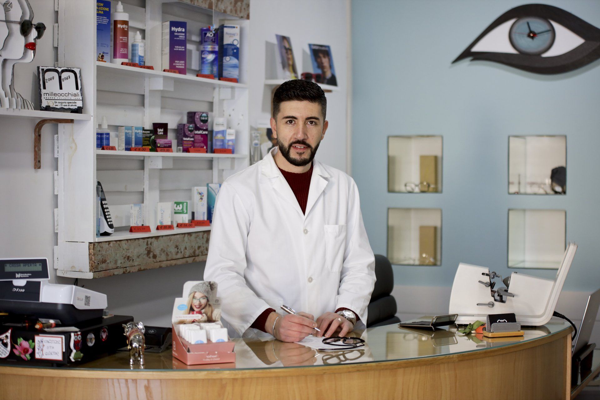Un ottico in camice bianco alla reception guarda la telecamera in una clinica oculistica dalle pareti blu.
