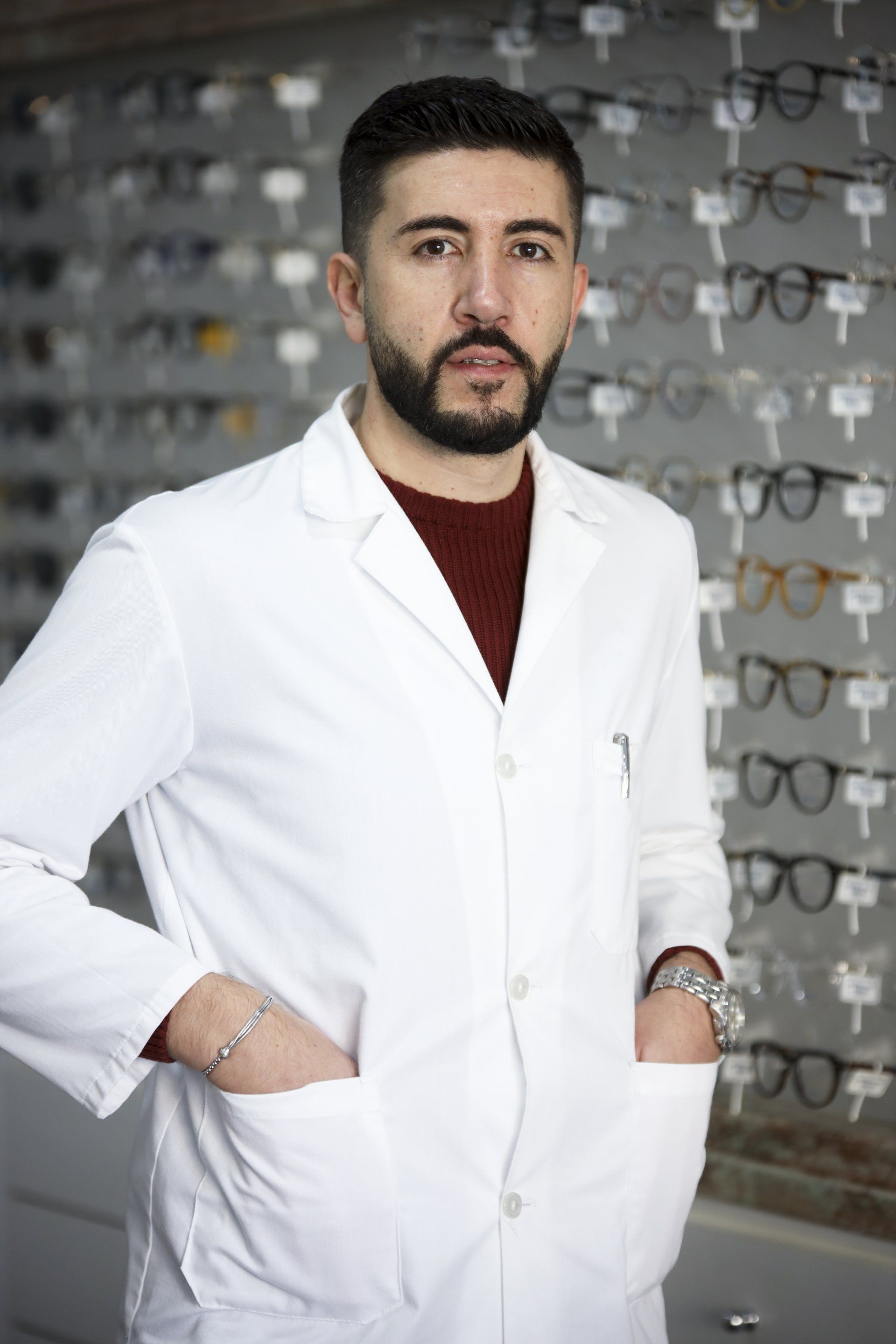 Un uomo in camice bianco, in piedi in un negozio di occhiali, con le mani in tasca, guarda la telecamera.
