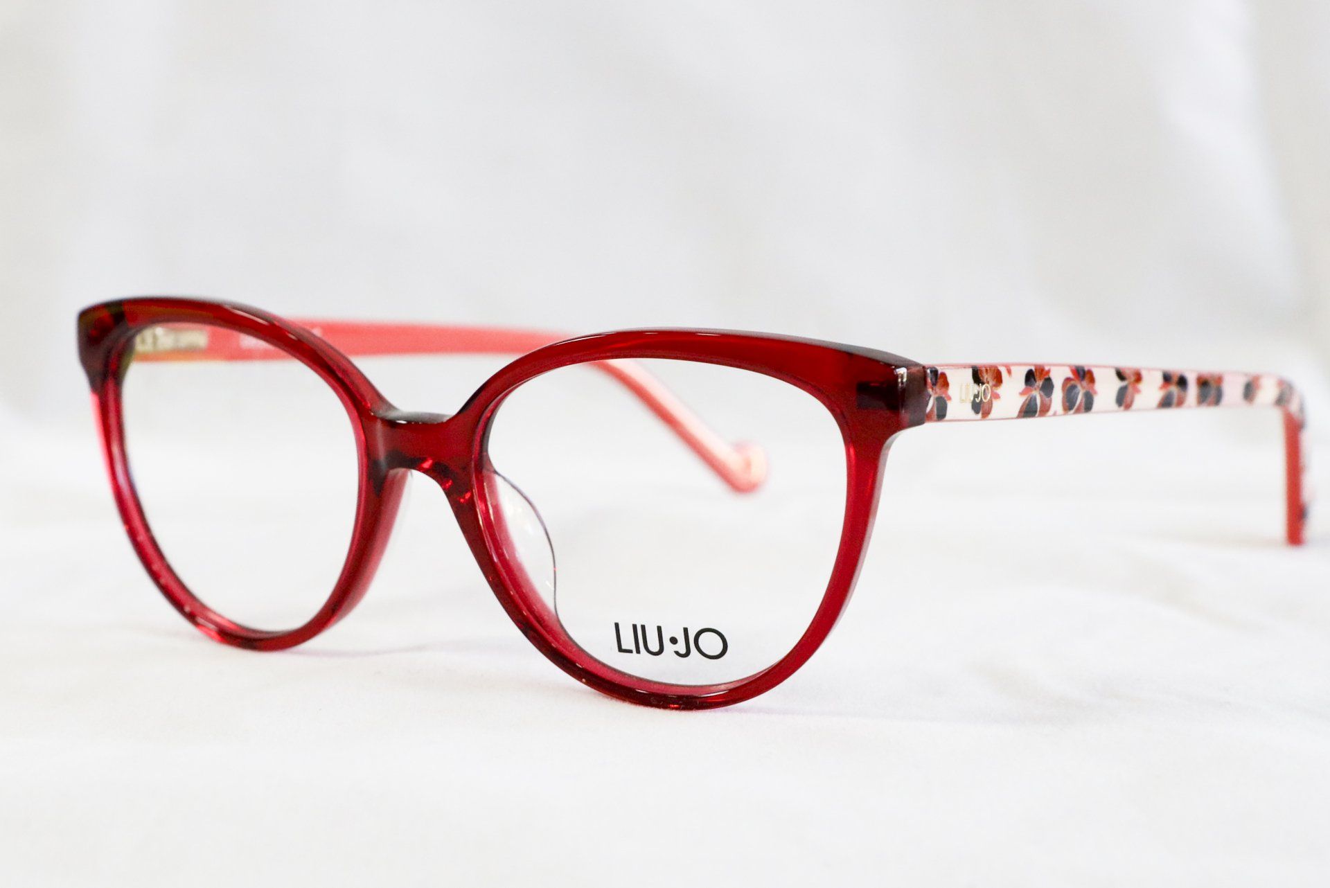 Occhiali da vista con montatura rossa e motivo floreale sulle aste, su sfondo bianco. Sulla montatura è stampata la scritta