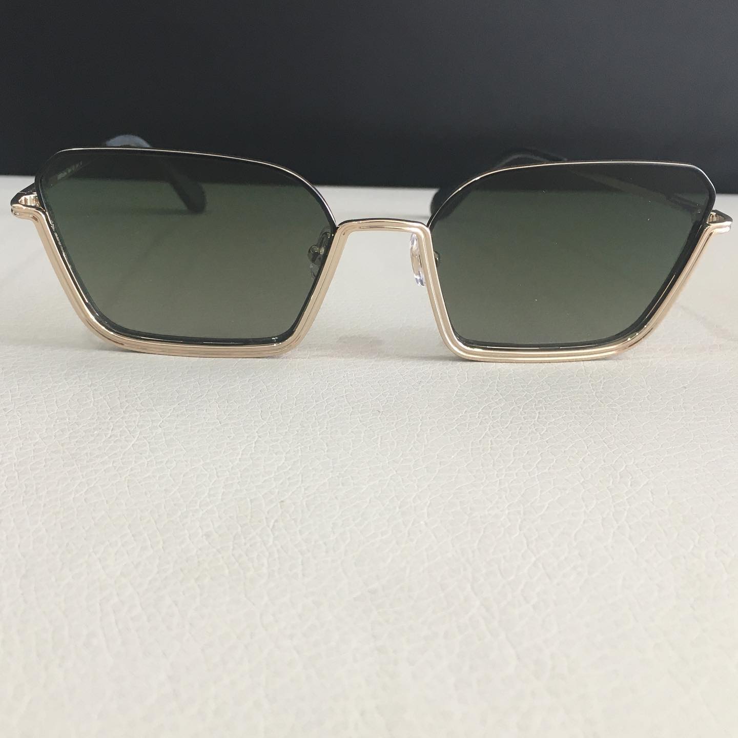 Occhiali da sole geometrici con montatura dorata e lenti verde scuro sono appoggiati su una superficie bianca, su uno sfondo scuro.