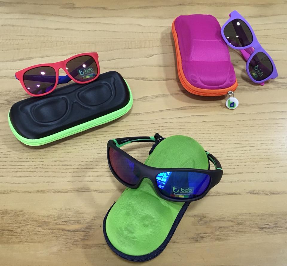 Occhiali da sole esposti su custodie: montature rosse, viola e nere. Le custodie sono verdi, rosa e nere. Superficie in legno.