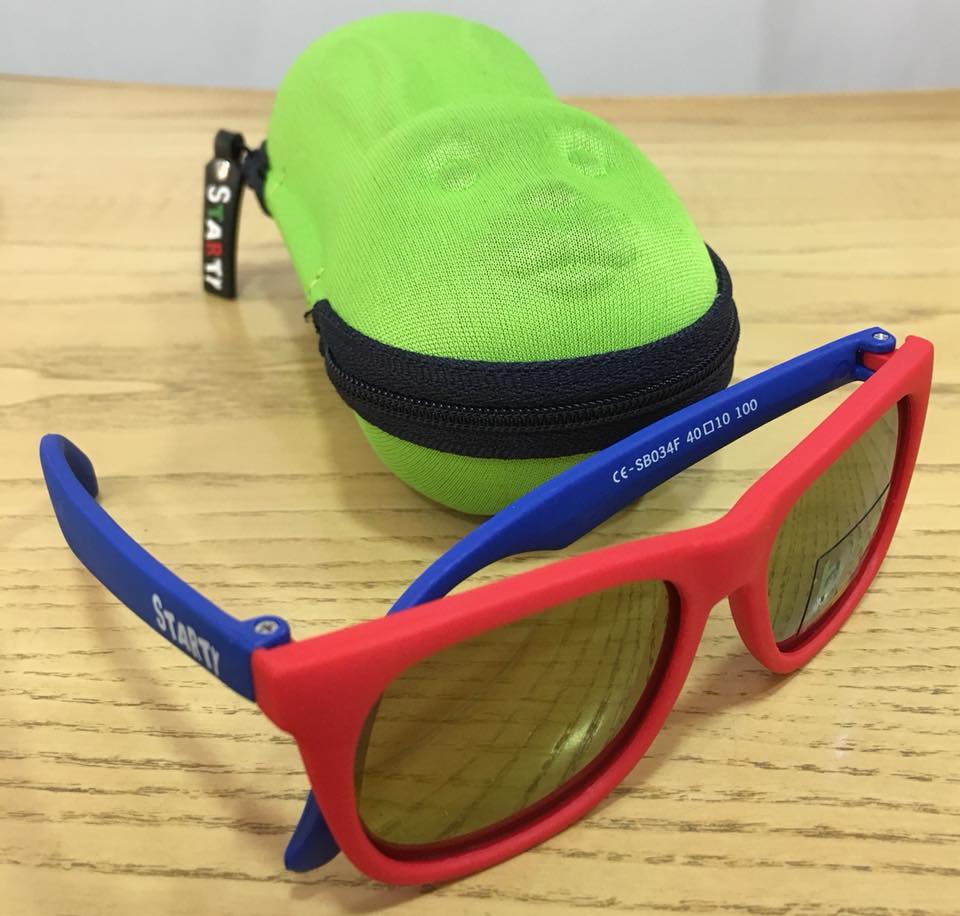 Occhiali da sole con montatura rossa e stanghette blu accanto a una custodia verde per occhiali da sole su una superficie di legno.