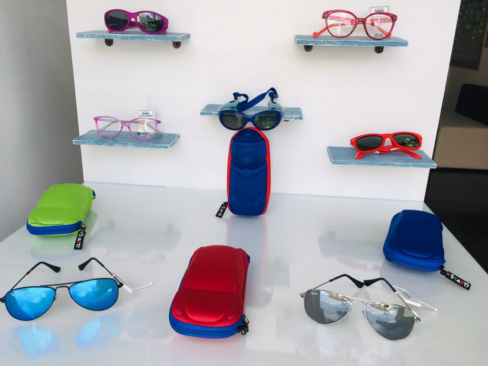 Occhiali da sole esposti sugli scaffali e su un bancone, in vari colori tra cui blu, rosso e rosa.