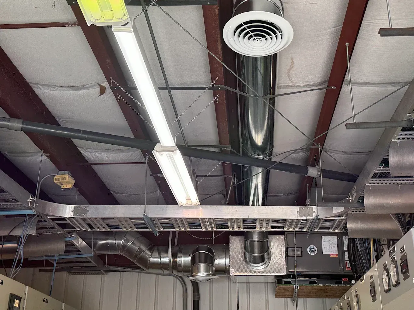 Industrial ceiling with ventilation, lights, and wiring. Metal ducts and beams against a white and brown backdrop.
